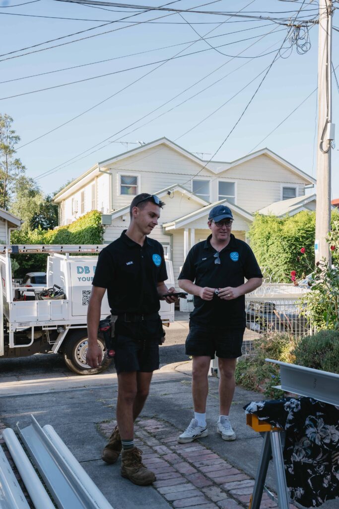 Two DB Pro Plumbing workers talking on a residential driveway in front of a branded work truck, holding tools and wearing uniforms. DB Pro Plumbing.